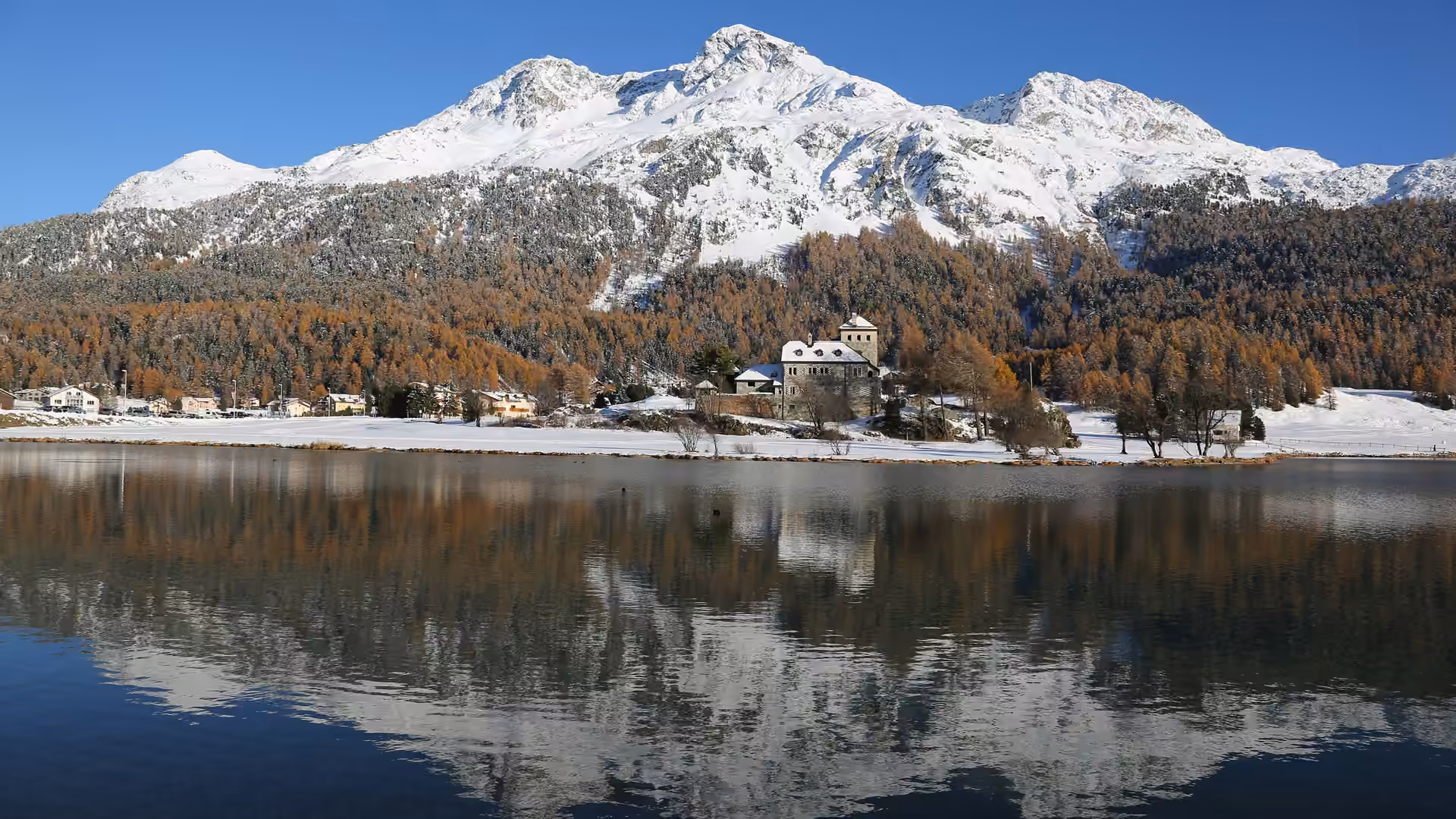 Azure Skies Resort za soumraku nad jezerem Silvaplana, na vrcholcích v pozadí leží sníh.